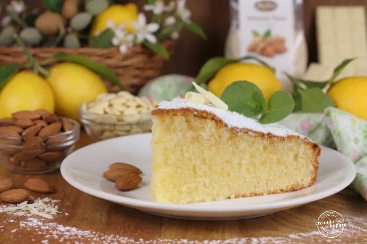 Una fetta di soffice torta caprese al limone, guarnita con zucchero a velo e foglioline di menta, circondata da limoni freschi e mandorle.