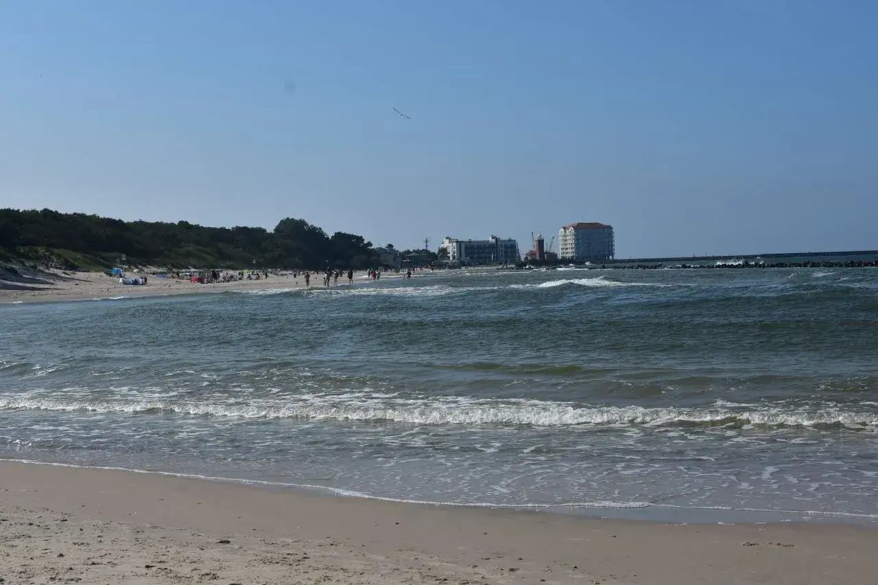 Słoneczny dzień nad Bałtykiem, plaża pełna ludzi, a w oddali widać miasto u ujścia Wieprzy.