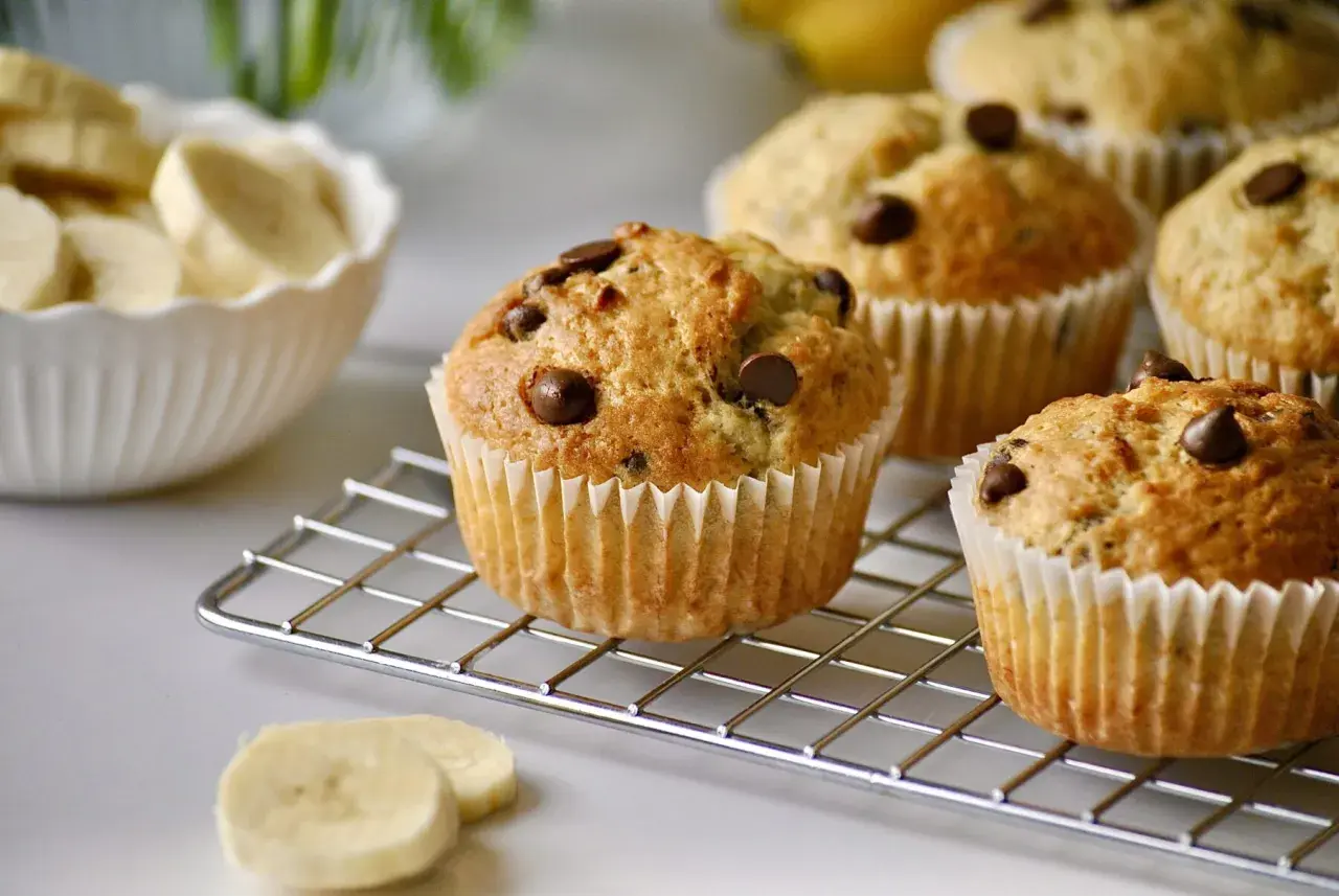 Frisch gebackene schoko bananen muffins mit Schokostückchen auf einem Kuchengitter. Daneben eine Schüssel mit Bananenscheiben.