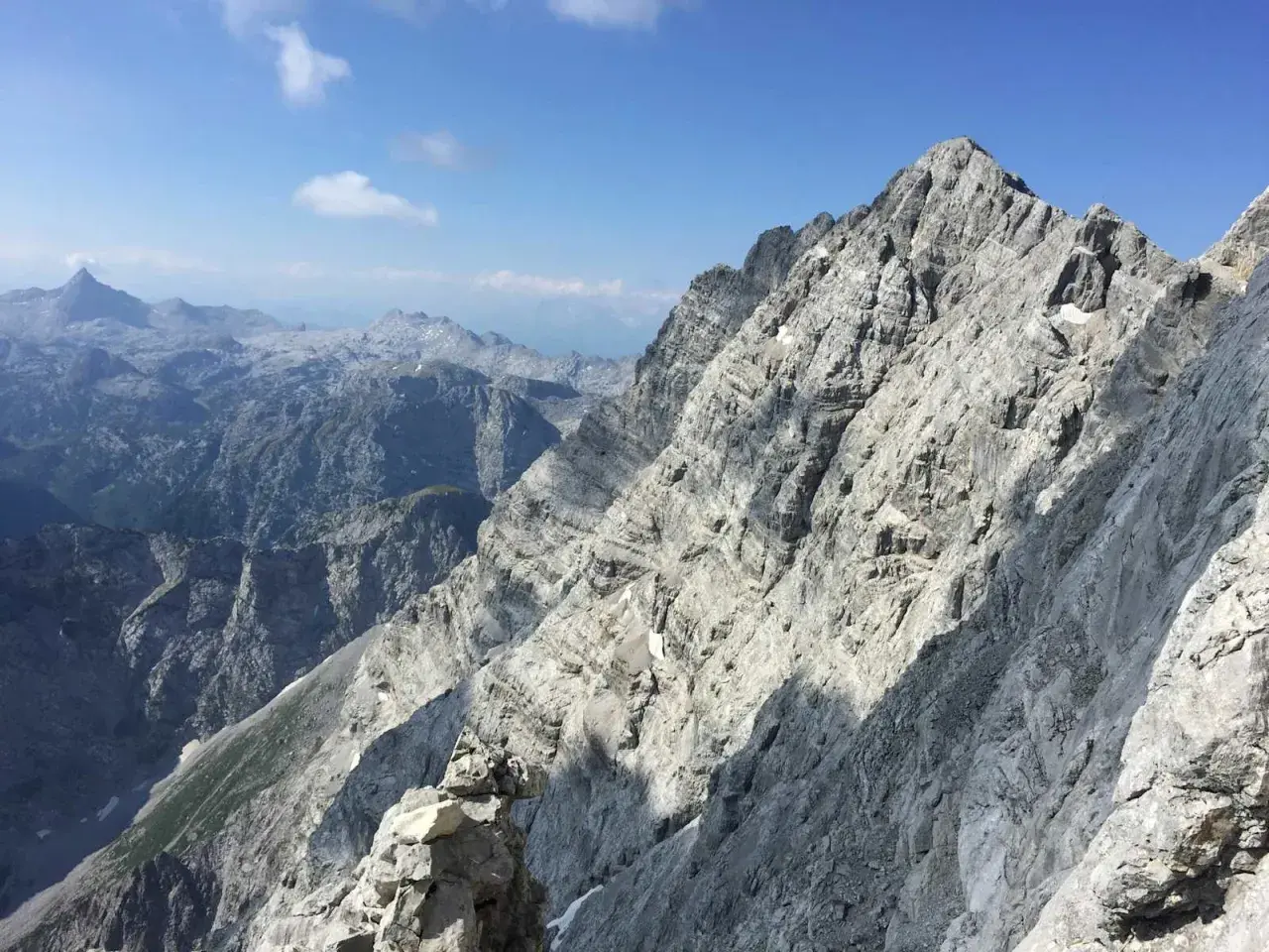 Steile Felsformationen der Watzmann Ostwand im Sonnenschein, mit tiefen Tälern und weiteren Berggipfeln in der Ferne.
