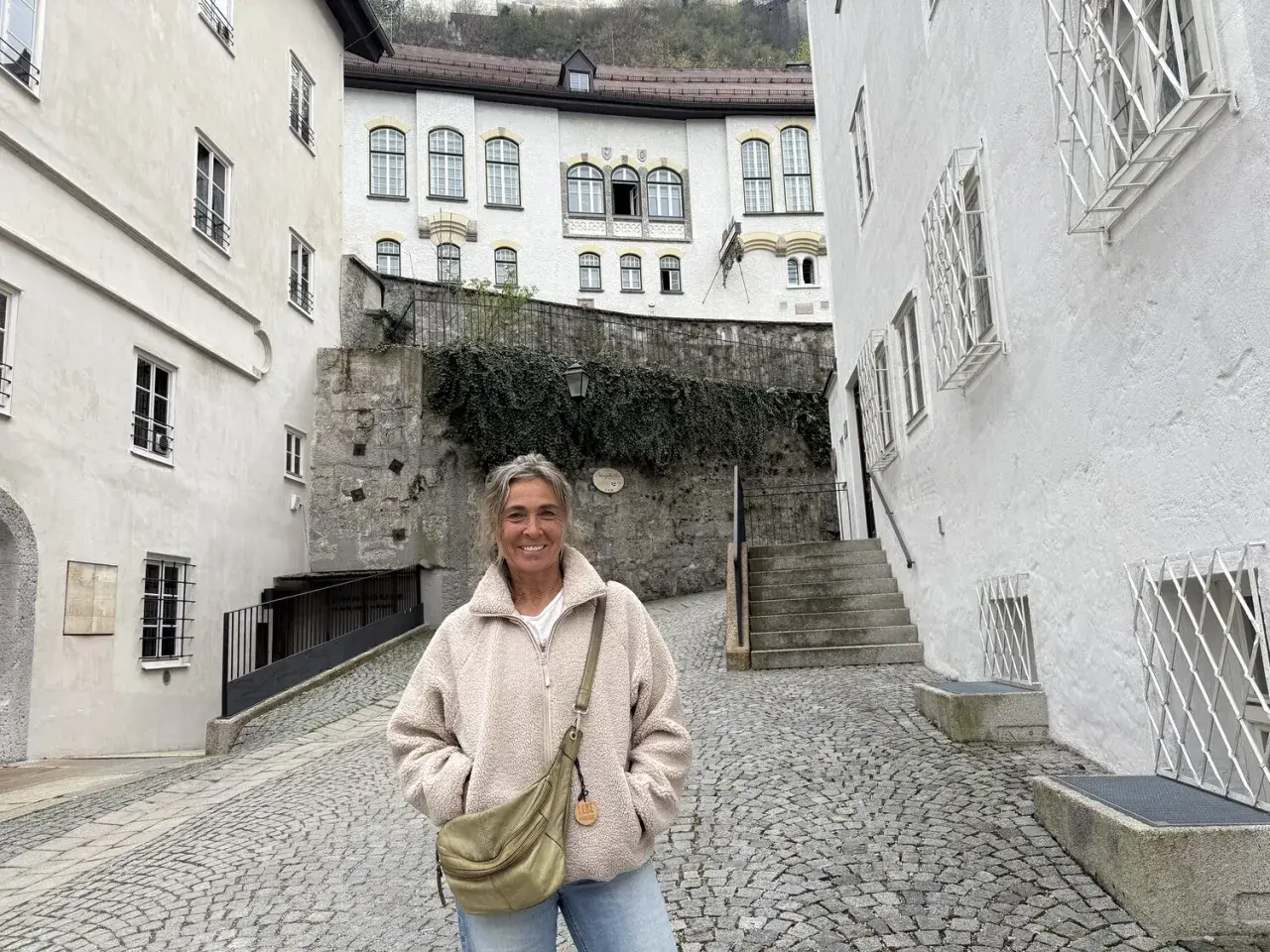 Eine Frau steht lächelnd auf einem Kopfsteinpflasterweg zwischen alten weißen Häusern. Sie könnte eine Pilgerin sein, die auf ihrer Reise neue Orte entdeckt.
