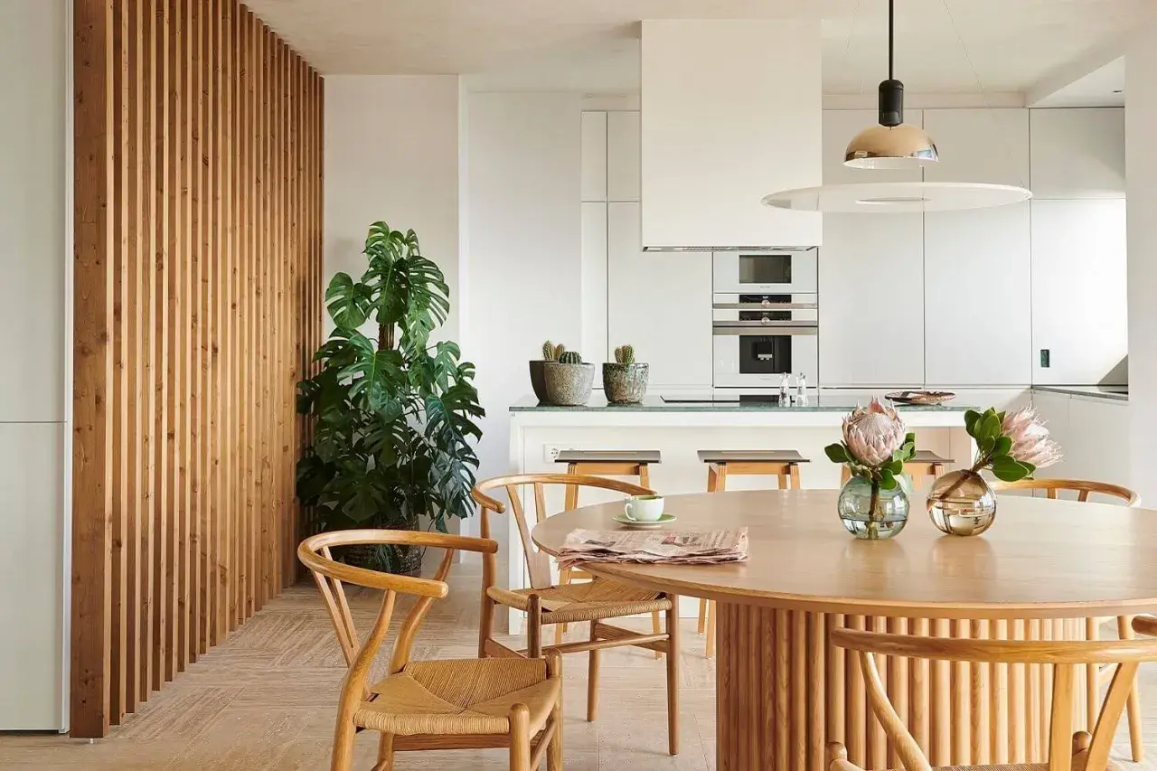 Comedor y cocina modernos con pared de lamas de madera, planta grande, mesa redonda y dos lámparas de techo.