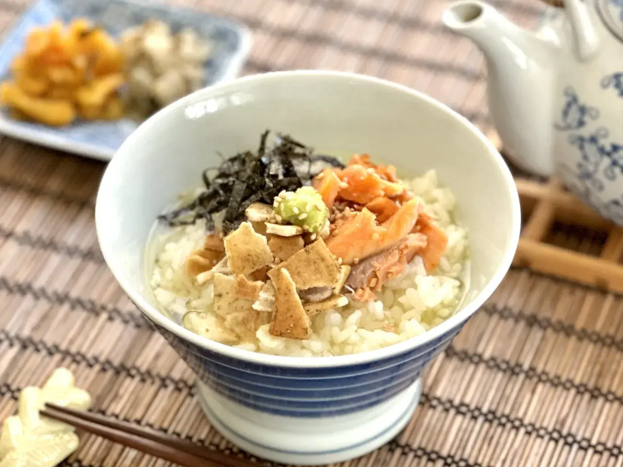 A delicious ochazuke recipe featuring rice, flaked salmon, nori, and wasabi in a blue-striped bowl, with a teapot in the background.