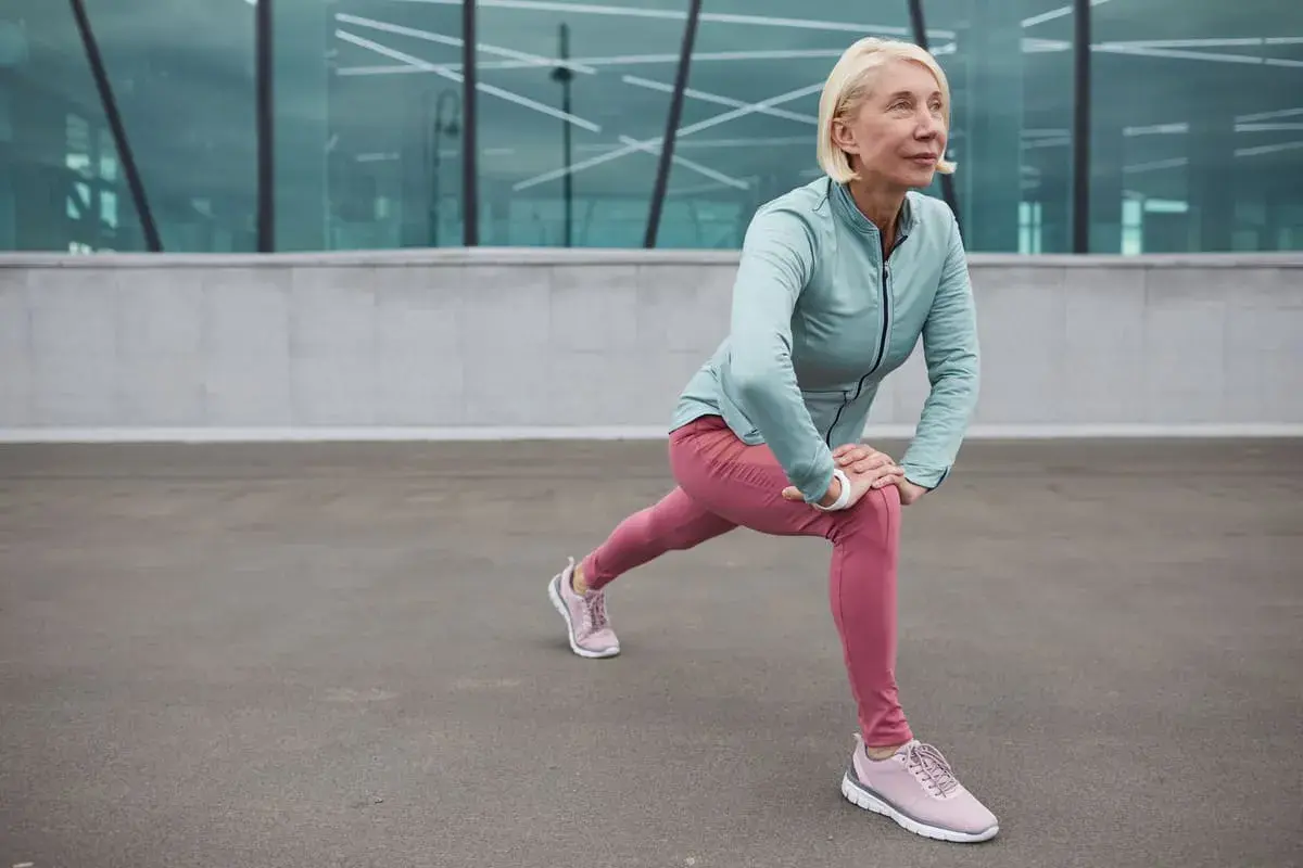 Donna bionda in abiti sportivi fa stretching, contrastando l'atonia muscolare con un affondo.