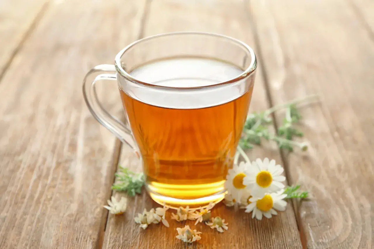 Ein Glas mit goldenem Kamillentee steht auf Holz. Kleine Kamillenblüten schmücken die Szene. Perfekt, um kamillentee kalt trinken.