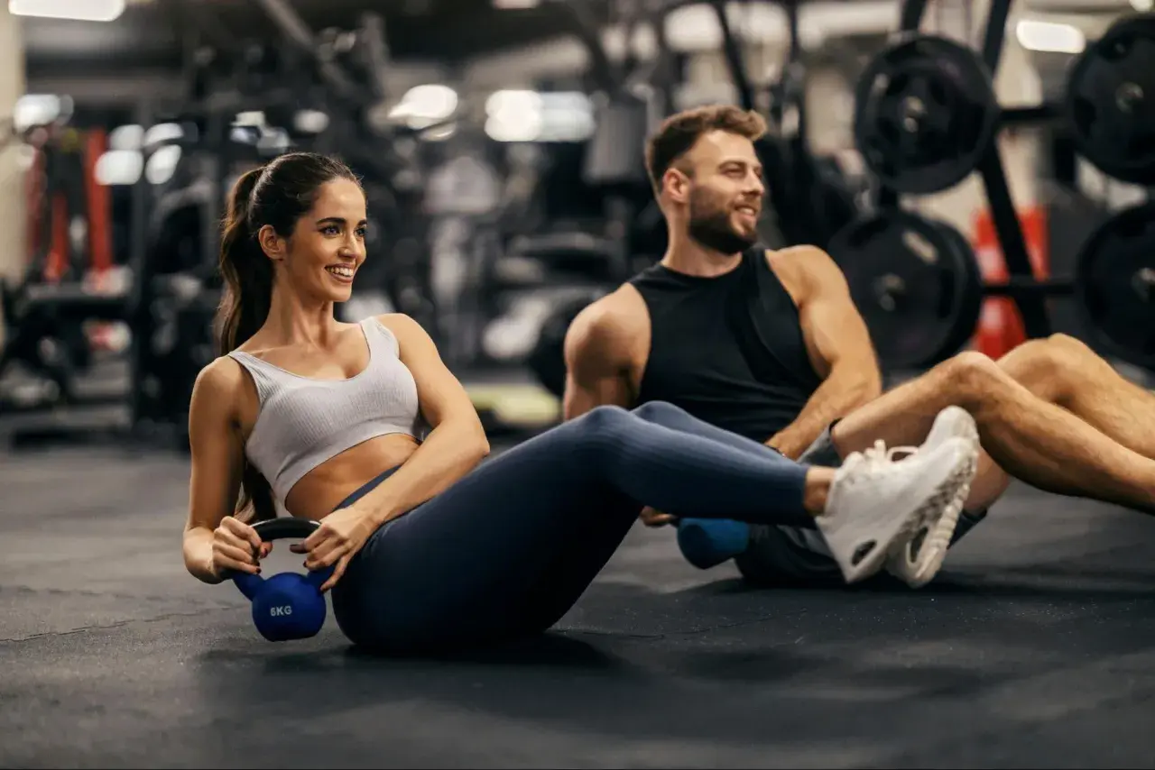 Couple working out in a gym, doing Russian twists with a kettlebell. This is part of how to train for an Ironman.