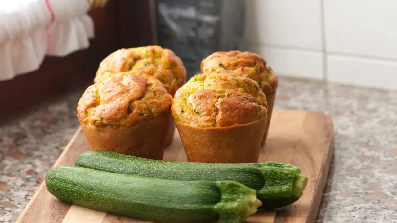 Vier goldbraune Zucchini-Muffins auf einem Holzbrett, daneben zwei frische Zucchini.