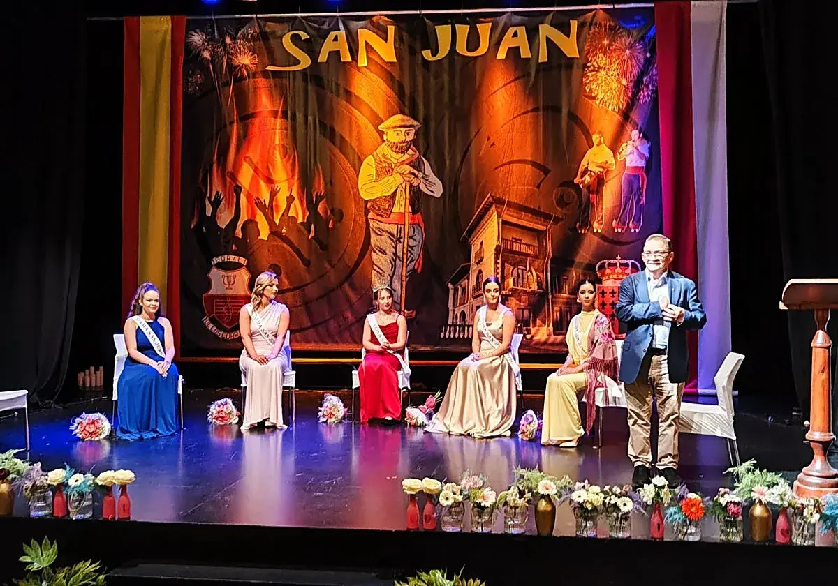 En el teatro Los Corrales de Buelna, cinco damas de honor y un presentador esperan en el escenario bajo un telón de San Juan.