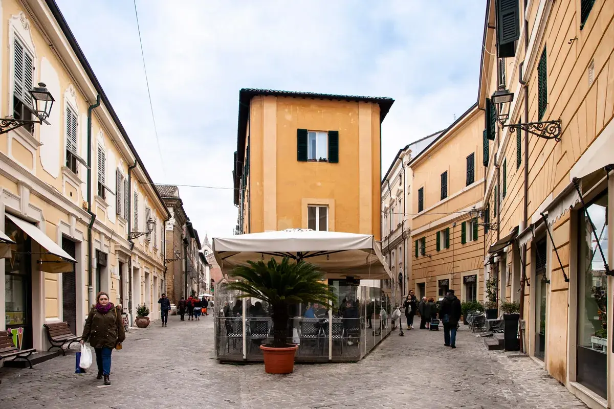 Una donna passeggia nel centro di Senigallia, tra palazzi color pastello e un dehors con palma.