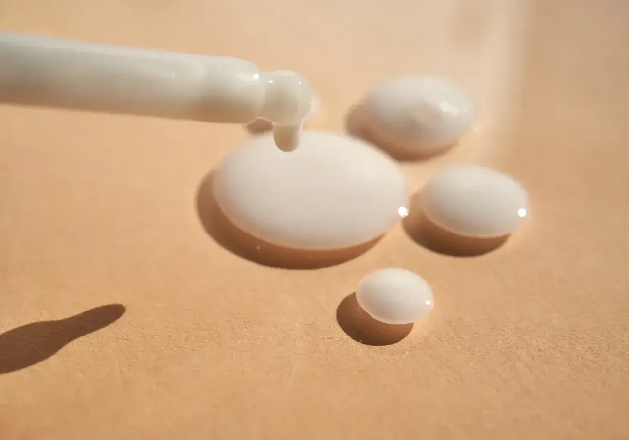 Ein weißes Serum tropft aus einem Applikator auf eine beige Oberfläche. Das ist Panthenol, ein Wirkstoff für Haut und Haar.