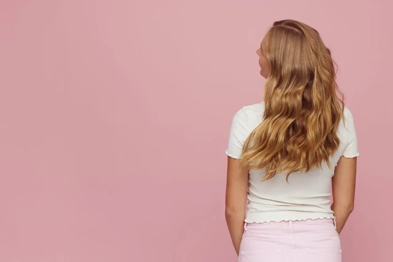 Blond włosy z efektem butterfly cut, falujące i lśniące, na tle pudrowego różu.