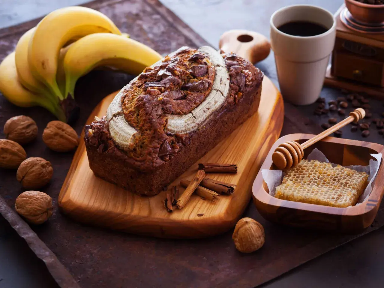 Ein saftiges Bananenbrot mit Schokoladenstückchen und Nüssen, garniert mit einer Banane. Daneben Kaffee, Honigwabe und Zimtstangen – ein Genuss, der auch bananenbrot gesund macht.