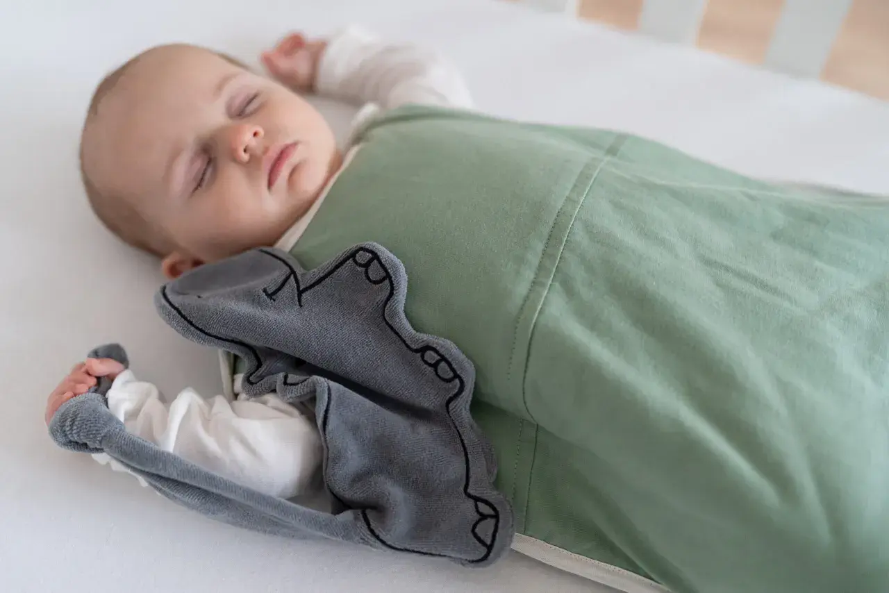 A baby sleeps soundly in a green sleep sack, a grey elephant toy peeking out. This image inspires thoughts on how to make baby clothes.