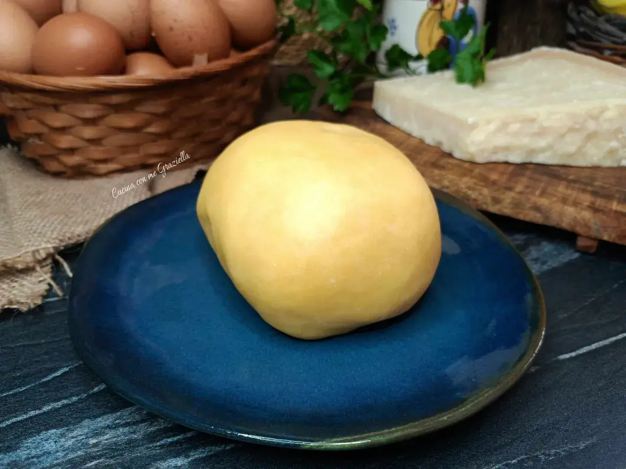 Impasto giallo per frolla salata su piatto blu, con uova e parmigiano sullo sfondo.