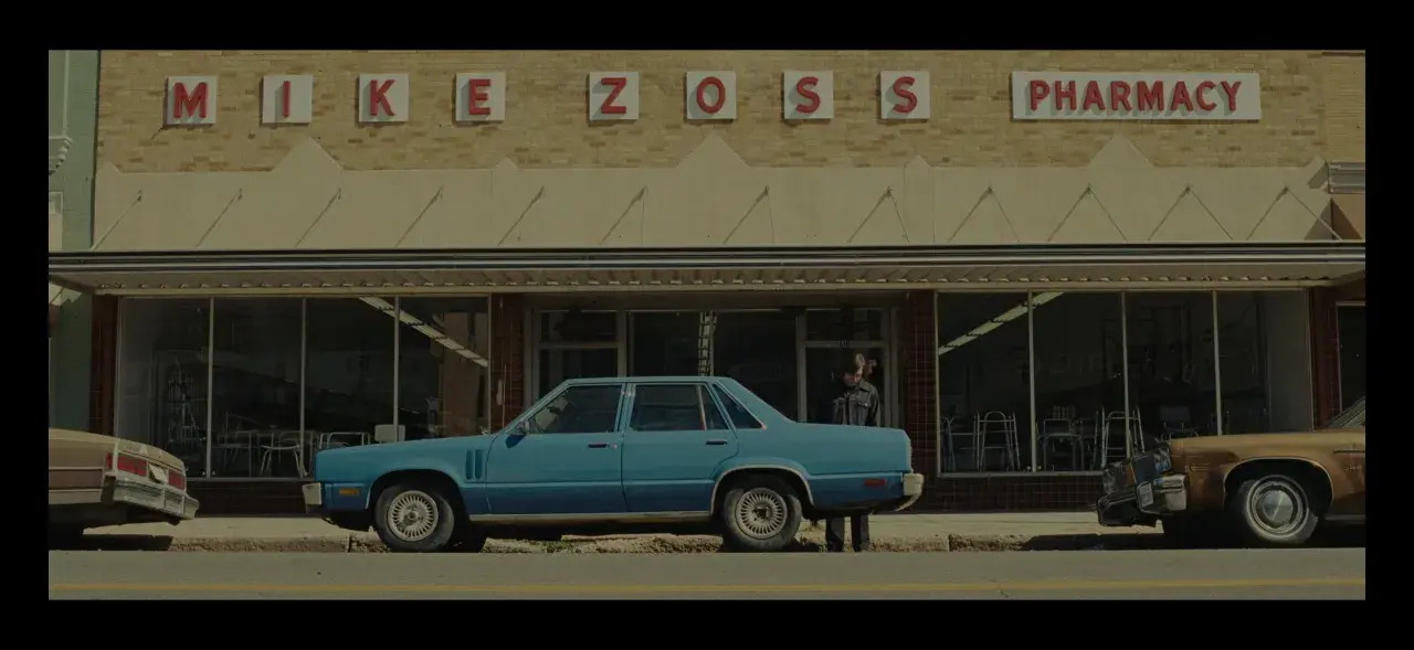 Niebieski samochód przed apteką "MIKE ZOSS PHARMACY". Obraz w stylu **false color**, z lekko zmienionymi barwami, tworzy nostalgiczny klimat.