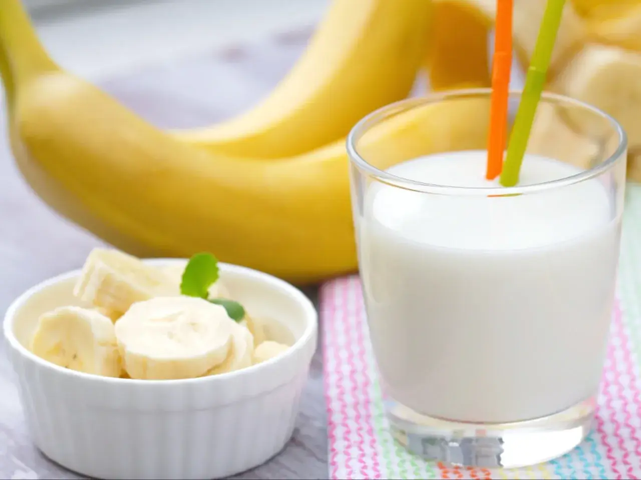 Szklanka pysznego mleka bananowego z dwoma słomkami, obok miseczka z pokrojonymi bananami i całe banany w tle.