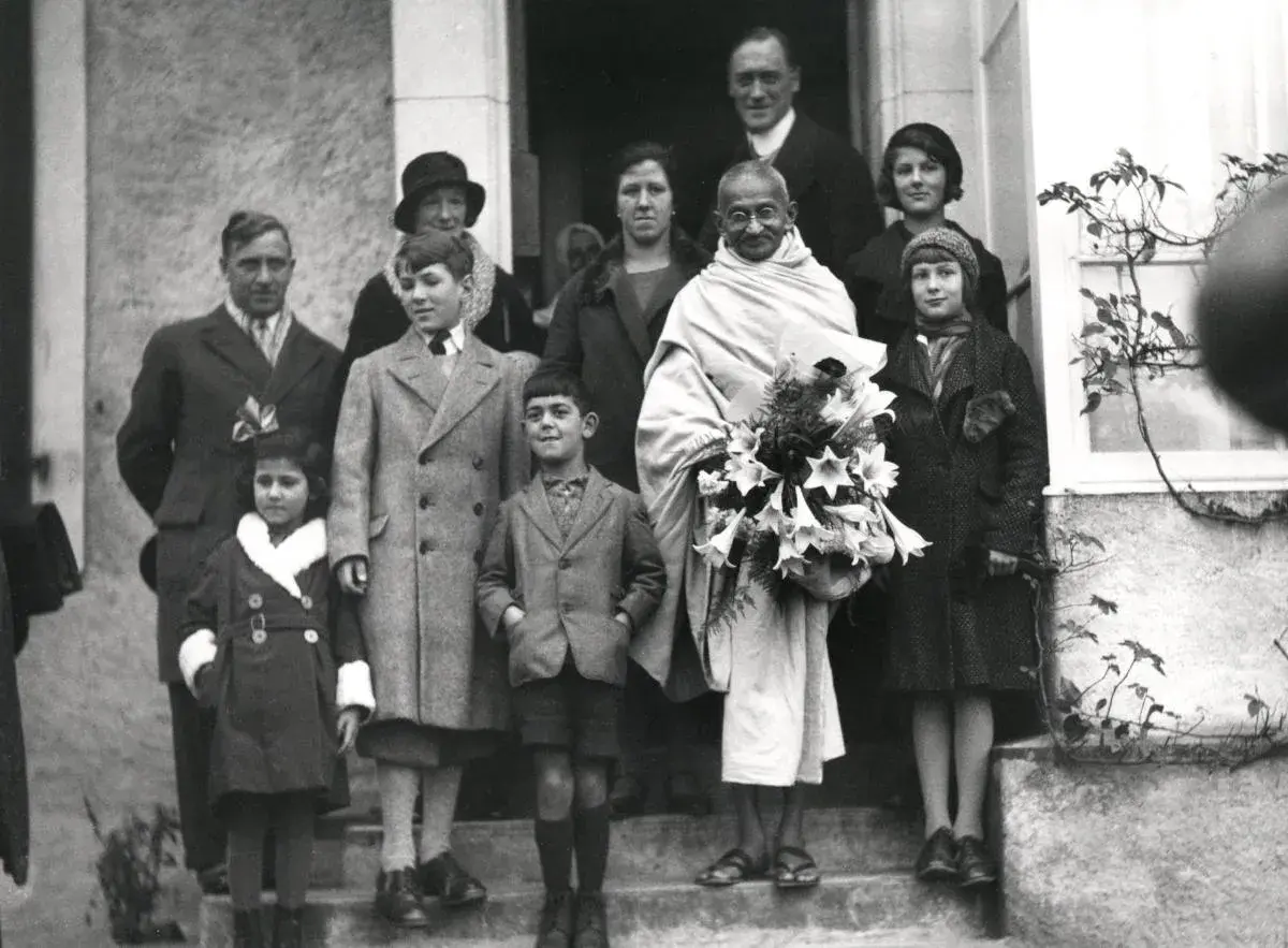Mahatma Gandhi mit seiner Familie und Freunden, umgeben von Kindern, die Blumen halten.