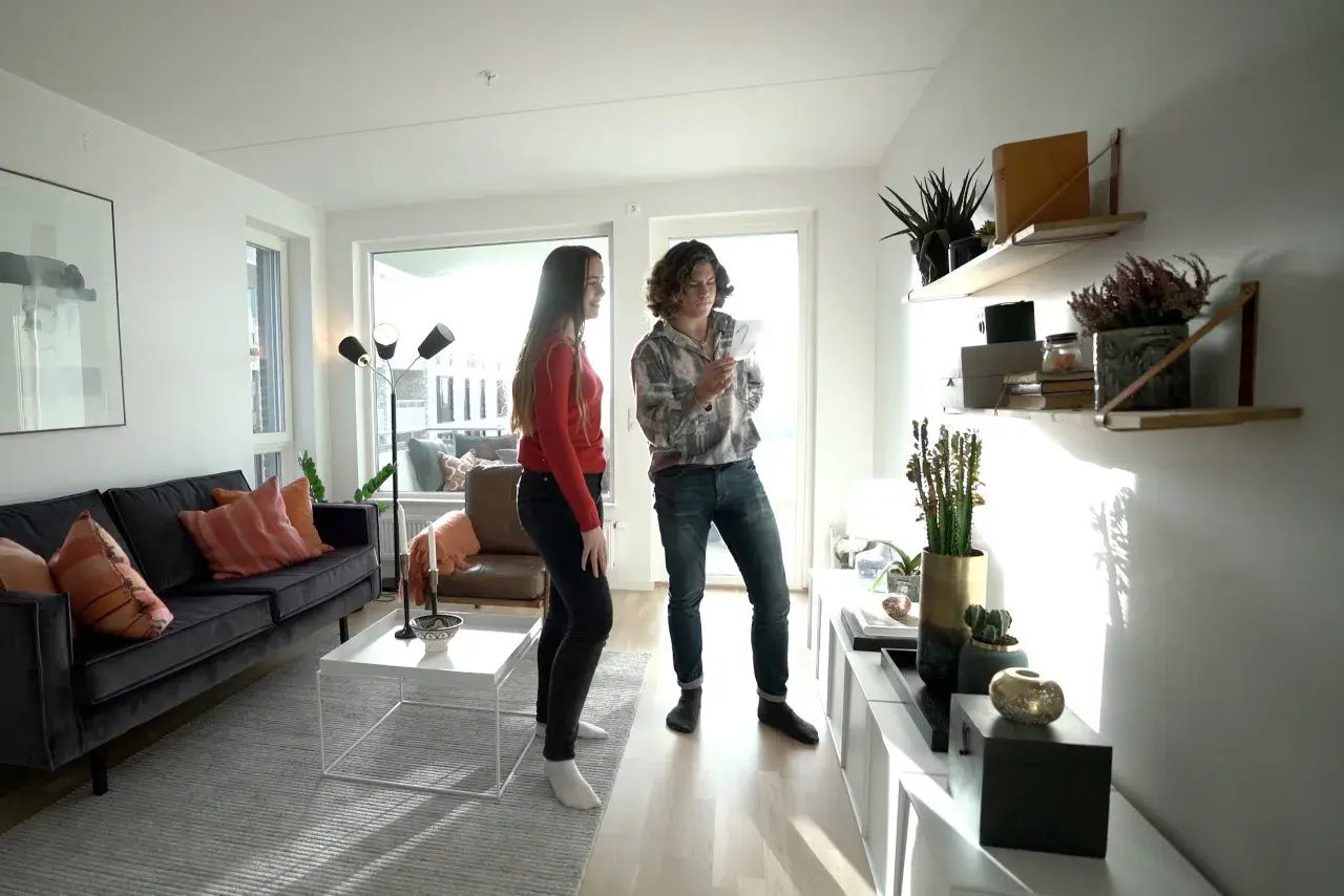 Två personer i ett ljust vardagsrum med stilren inredning. En mörk soffa med kuddar, ett vitt soffbord och en golvlampa. Hyllor med växter och dekorationer.