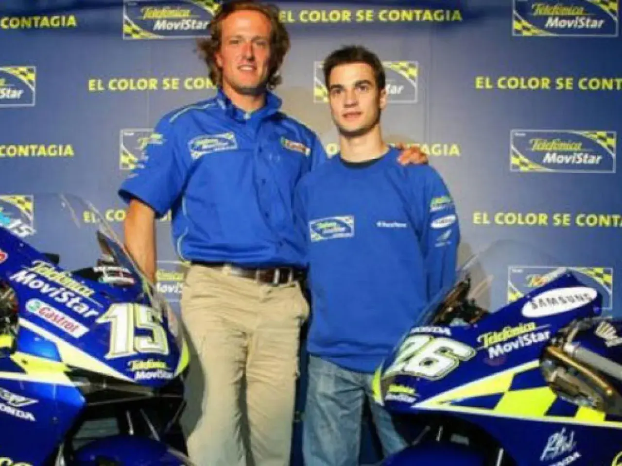 Sete Gibernau, con su edad, posa junto a un joven piloto, ambos con ropa de equipo azul, frente a motocicletas de competición.