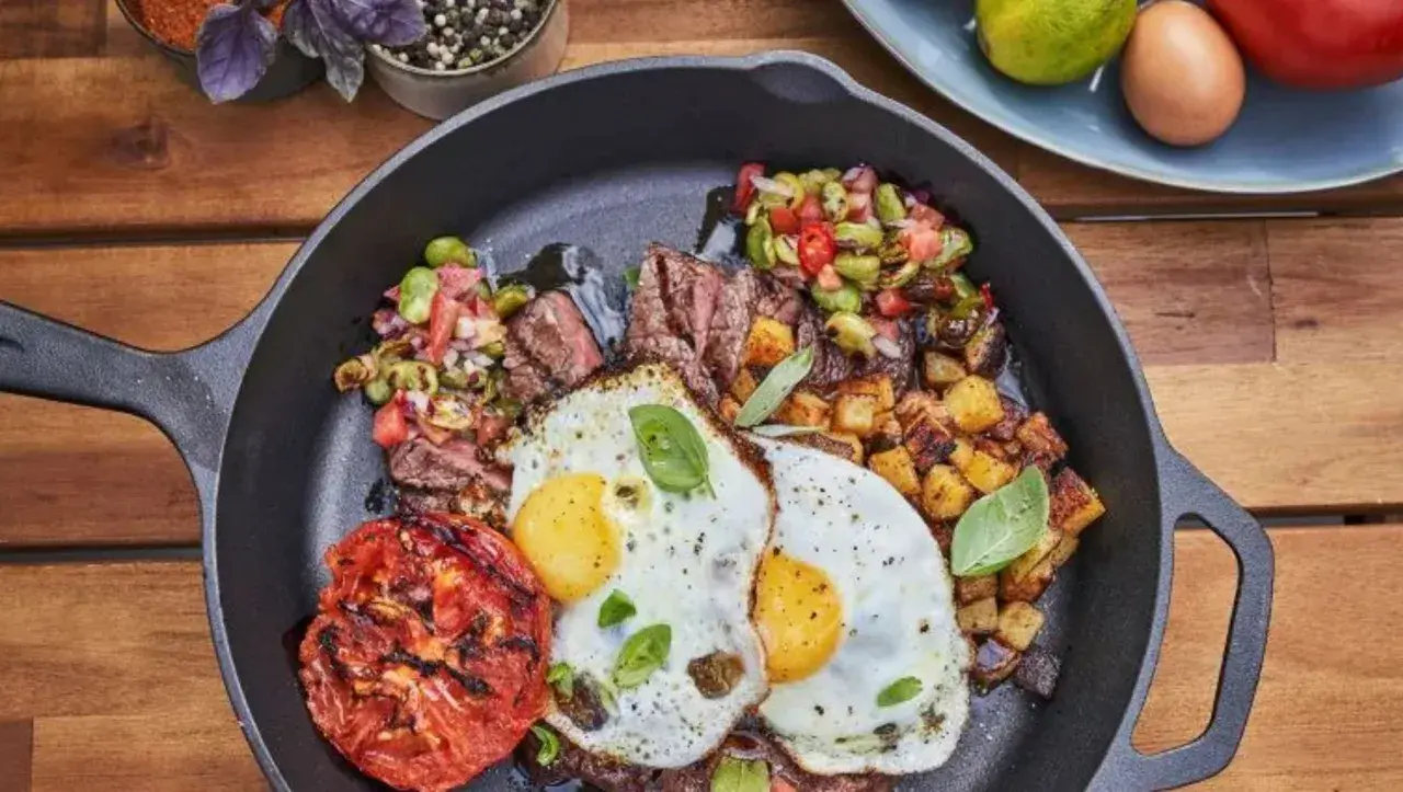 Un plat de petit-déjeuner copieux dans une poêle en fonte : steak grillé, deux œufs au plat, pommes de terre rôties, tomate grillée et une salade de légumes. Un vrai oeuf barbecue !