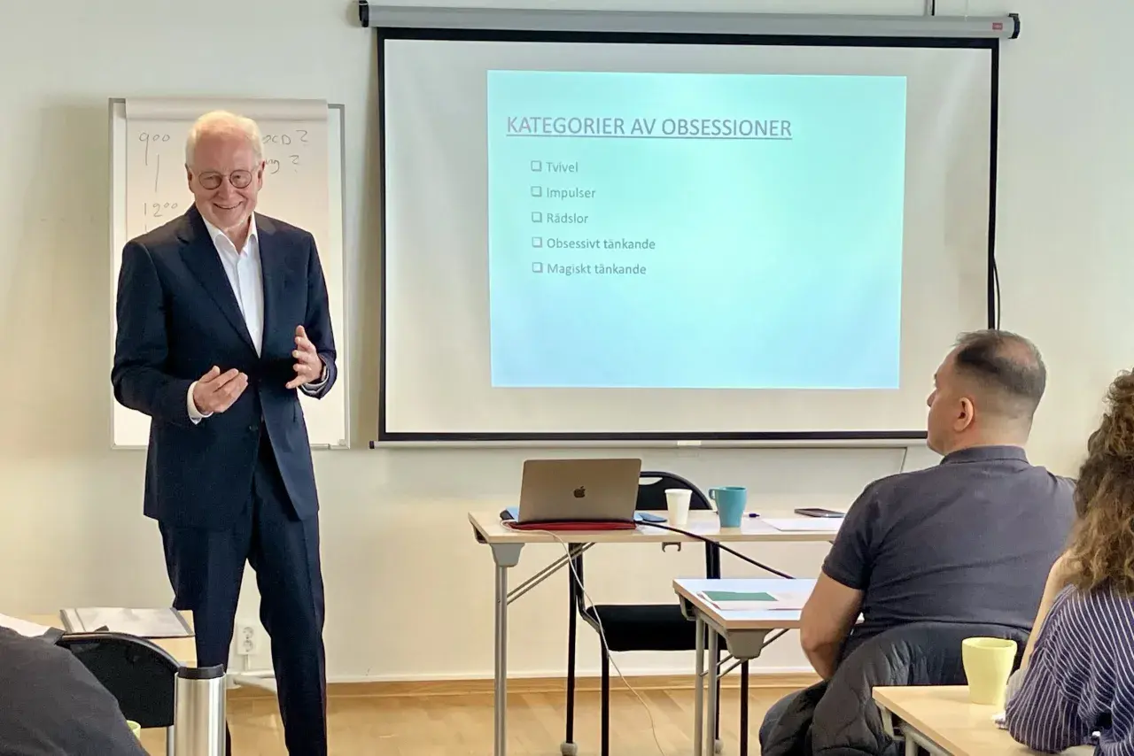 Föreläsare presenterar "Kategorier av obsessioner" som steg 1 i en utbildning. Publiken lyssnar uppmärksamt.