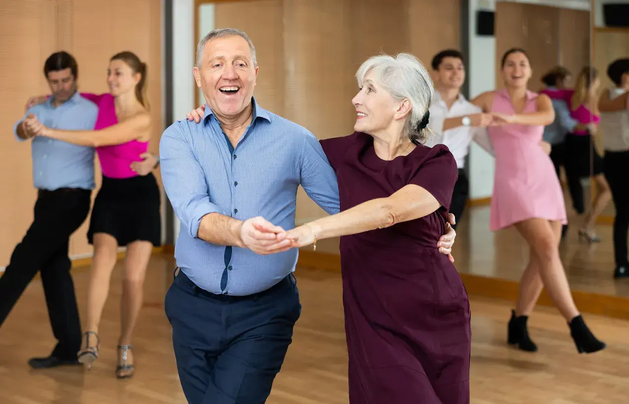 Uśmiechnięta para seniorów w rytmie tańca na 3. W tle inne pary ćwiczą kroki.