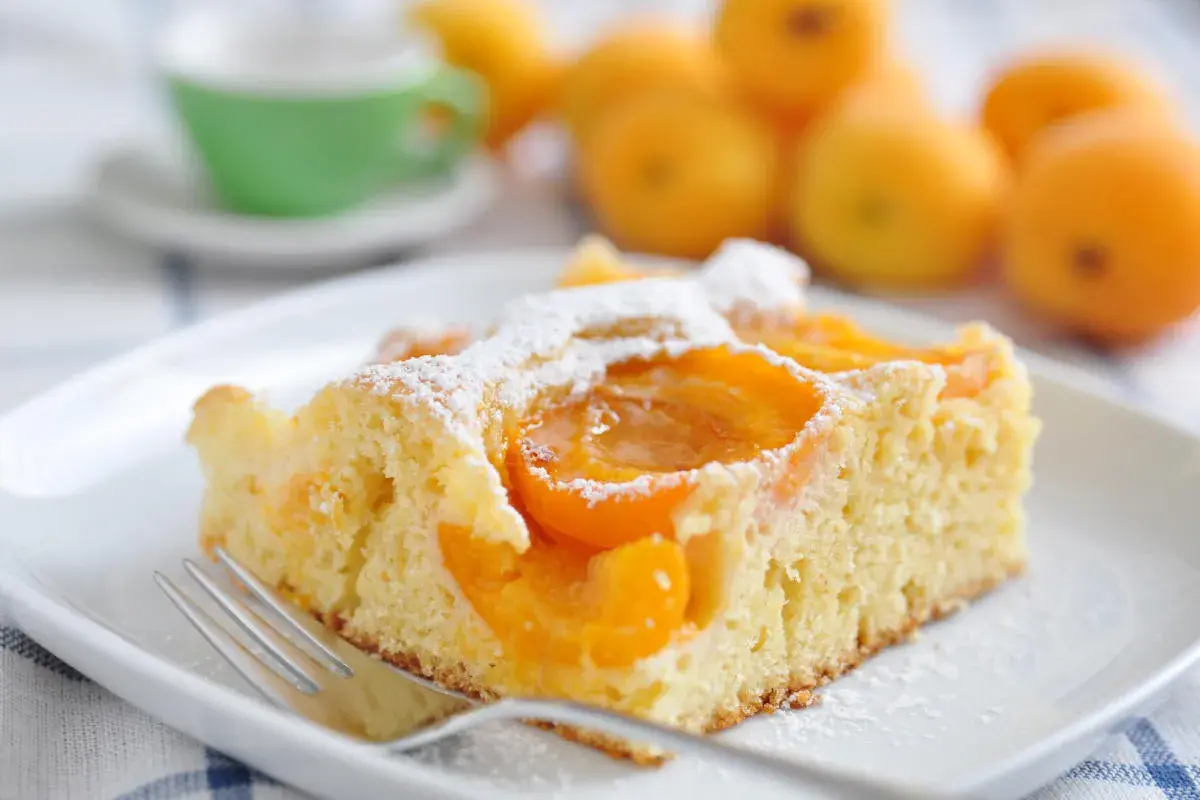 Una fetta di torta albicocche e yogurt, soffice e profumata, cosparsa di zucchero a velo. Sullo sfondo, albicocche fresche e una tazzina verde.