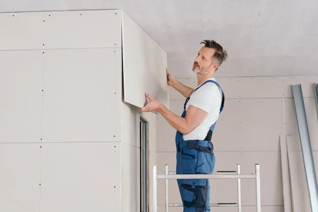 Operaio in tuta blu posiziona un pannello di cartongesso sul soffitto, completando il lavoro di fissare cartongesso direttamente su parete.