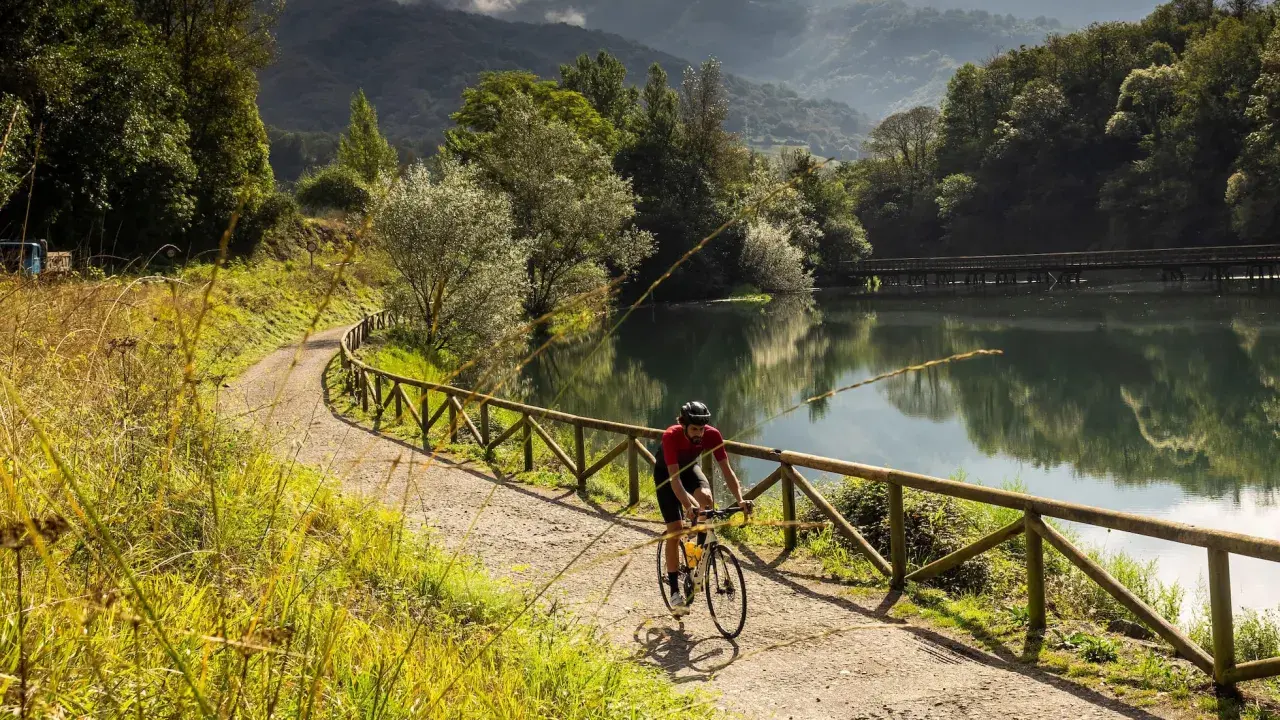 Ciclista pedalea por una de las vías verdes España, junto a un lago sereno y rodeado de exuberante vegetación.