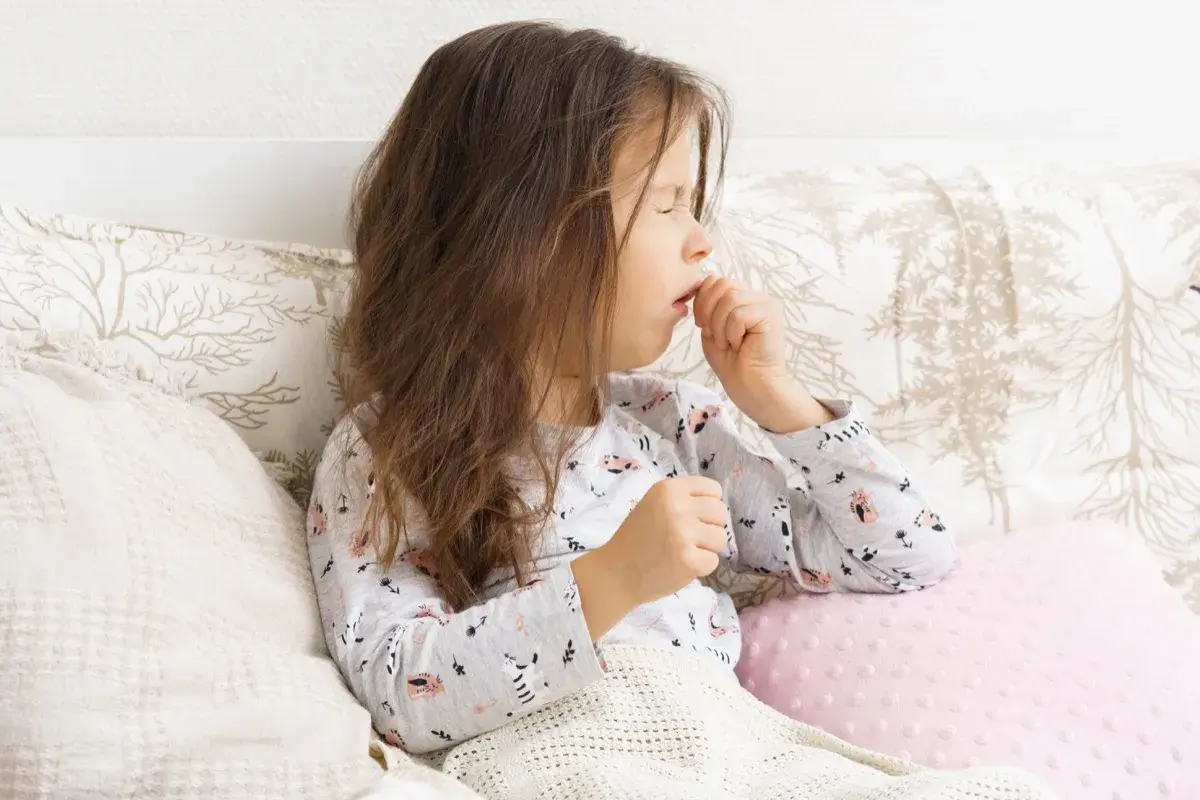 Bambina con tosse stizzosa, coperta da una coperta, seduta a letto con cuscini decorati.