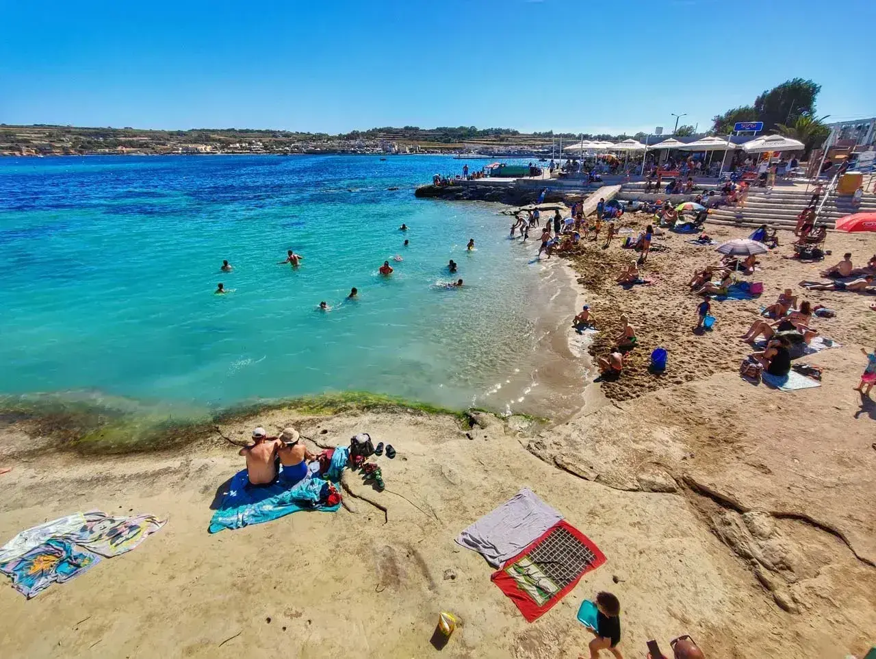 Piękna, słoneczna pogoda na Malcie. Ludzie kąpią się w turkusowej wodzie i odpoczywają na piaszczystej plaży pod parasolami.