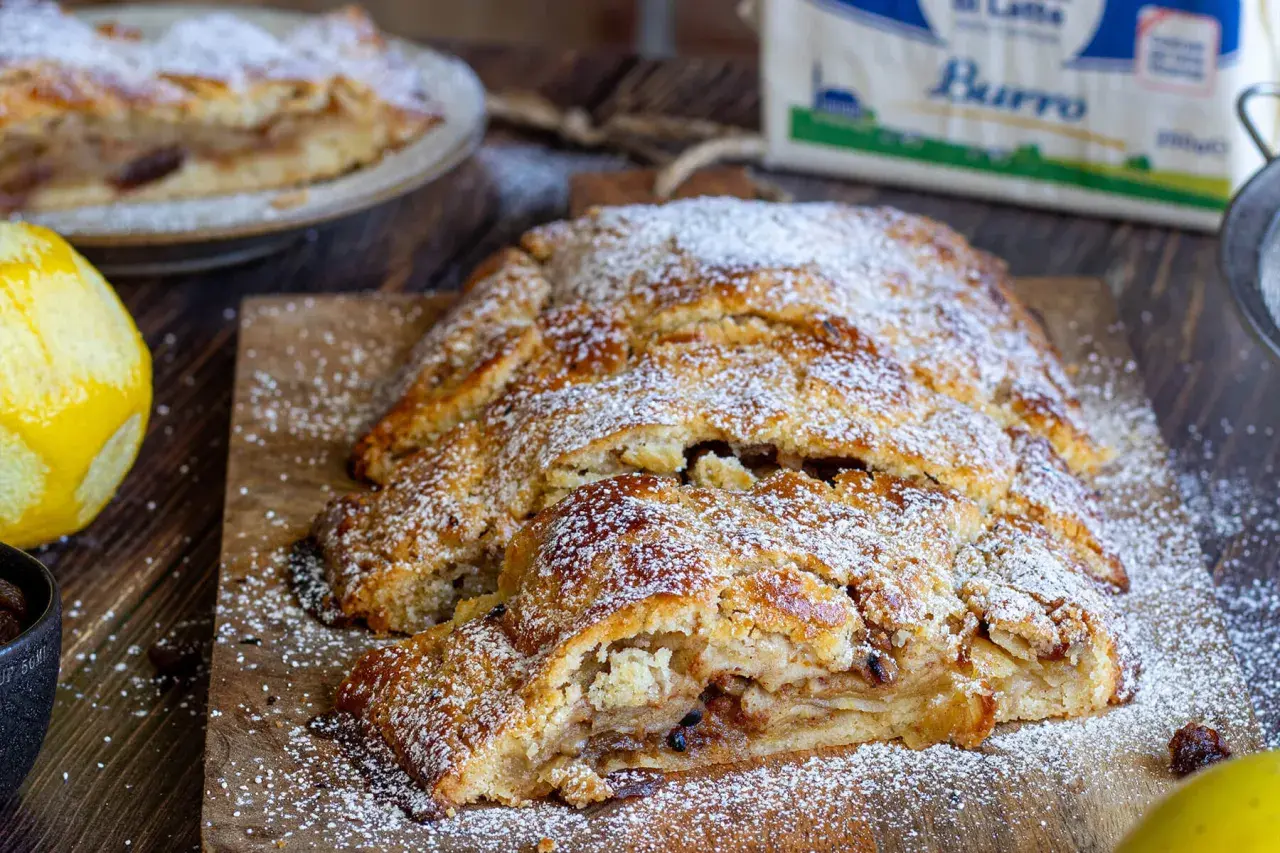 Strudel di pasta frolla dorato, cosparso di zucchero a velo, con ripieno di mele e uvetta. Accanto, scorza di limone e una tazza di uvetta.