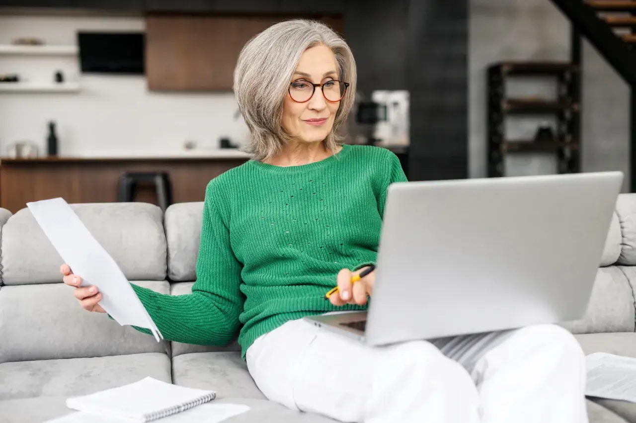 Femme âgée souriante, assise sur un canapé, travaillant sur son ordinateur portable et consultant des documents. Elle semble gérer son **apa à domicile montant**.