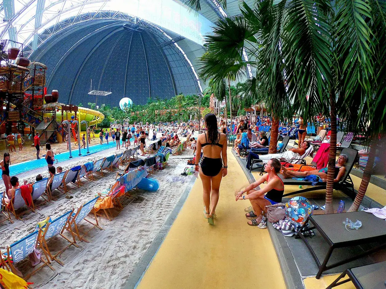 Tropikalny raj w aquaparku pod Berlinem. Ludzie odpoczywają na plaży pod ogromną kopułą, wśród palm i zjeżdżalni.