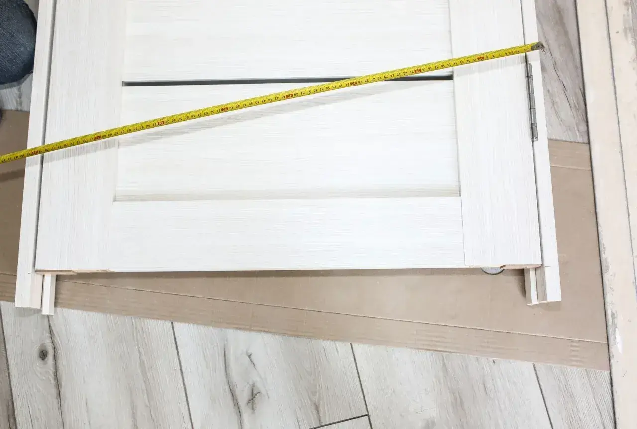 Białe drzwi z widoczną miarką. Mierzone skrzydło drzwiowe ma szerokość ok. 70 cm, co sugeruje jaki otwór na drzwi 70 będzie potrzebny.