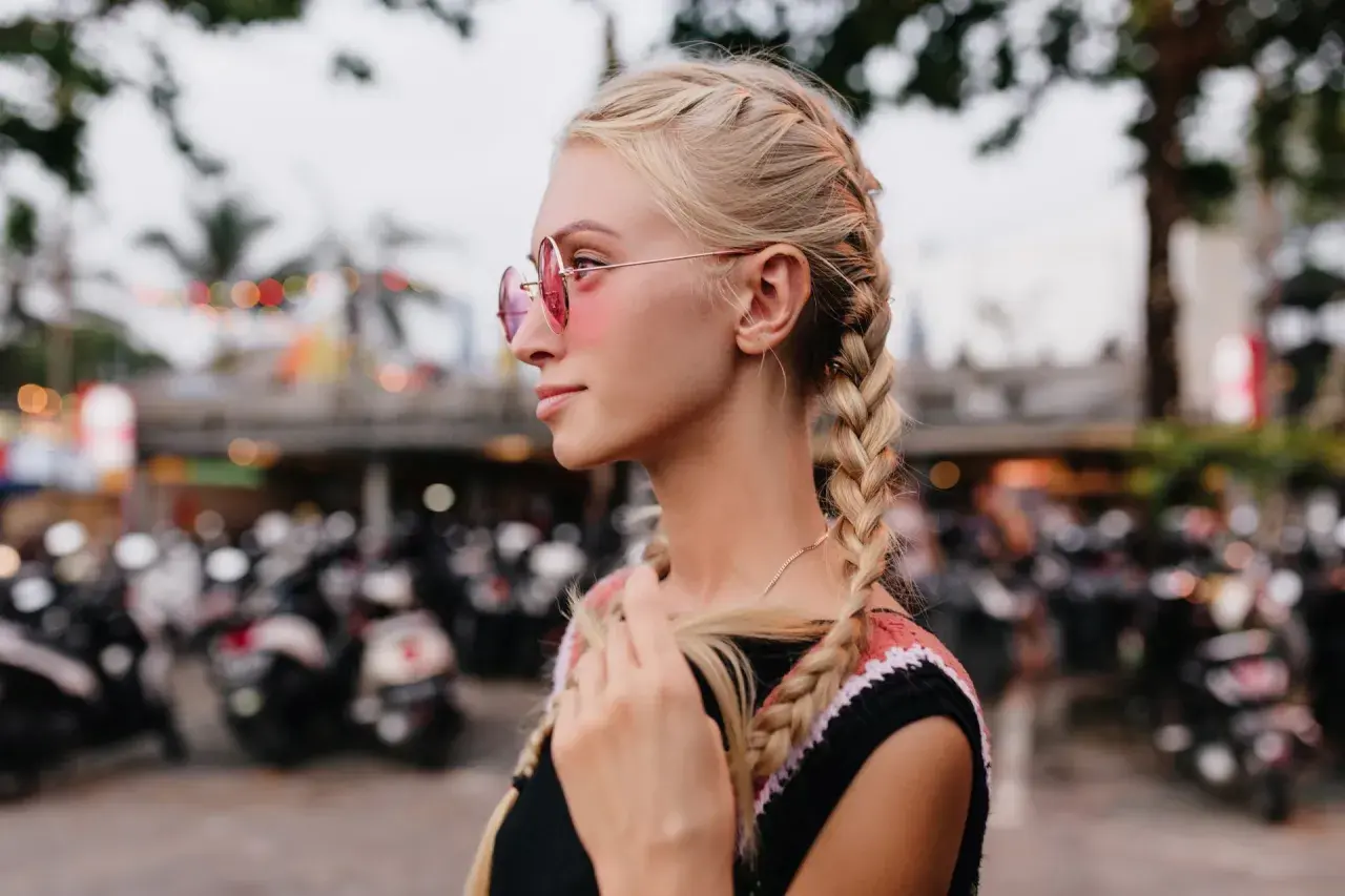 Blondynka w okularach przeciwsłonecznych z dwoma warkoczami na tle skuterów.