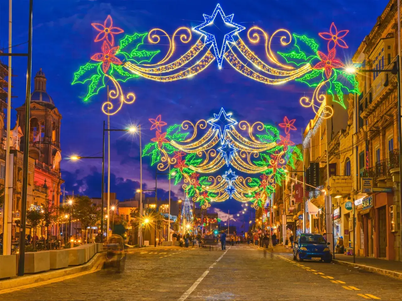 Malta w grudniu rozświetlona świątecznymi iluminacjami. Ulica ozdobiona girlandami z gwiazd i kwiatów, w tle kościół i sklepy.