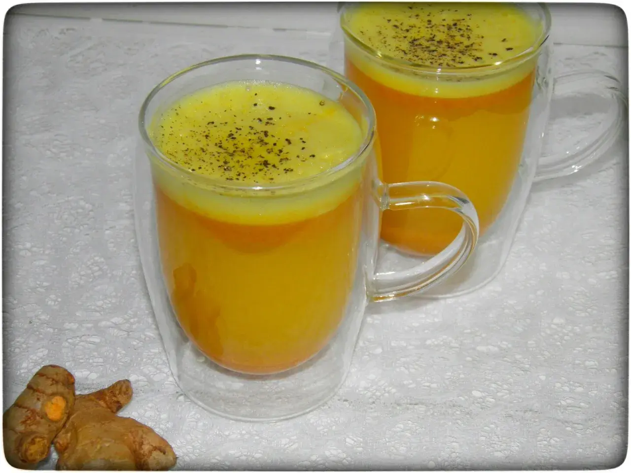 Zwei Gläser mit leuchtend gelbem Kurkuma-Wasser, bestreut mit schwarzem Pfeffer. Daneben liegen frische Kurkumawurzeln. Ein einfaches Rezept für Wohlbefinden.
