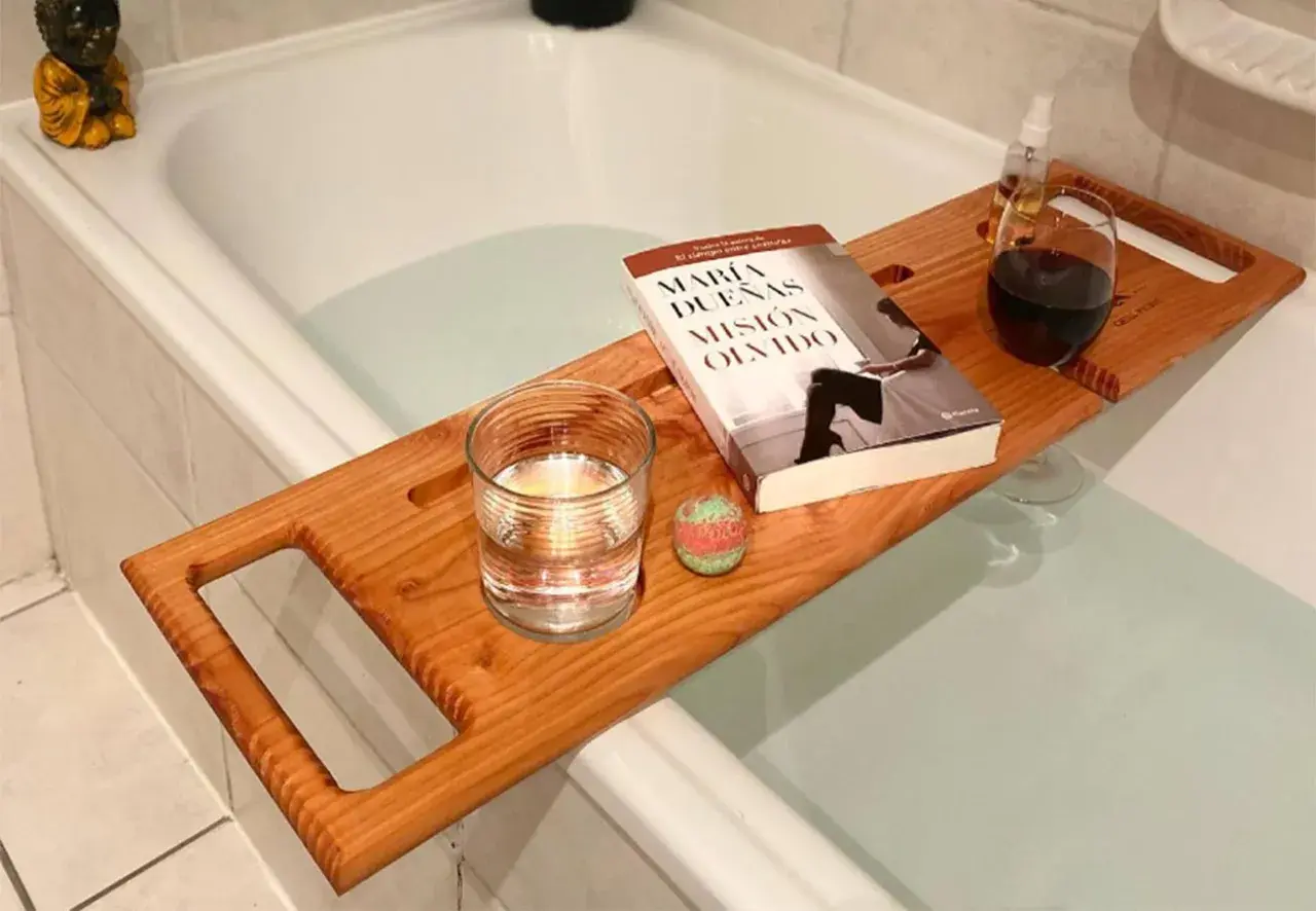 Tabla de madera sobre bañera con libro, vaso de agua, bomba de baño y copa de vino. Preparando un baño relajante.
