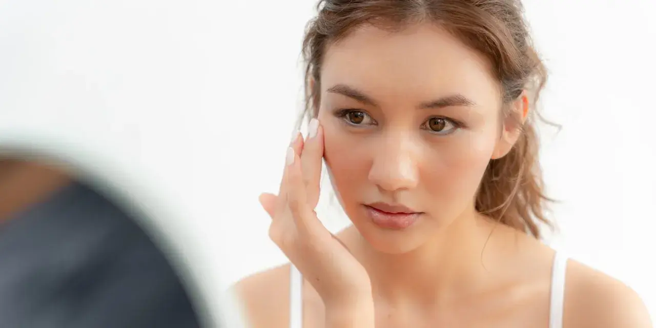 Une jeune femme examine son visage, préoccupée par la sécheresse cutanée. Sa peau semble tendue et manque d'hydratation.