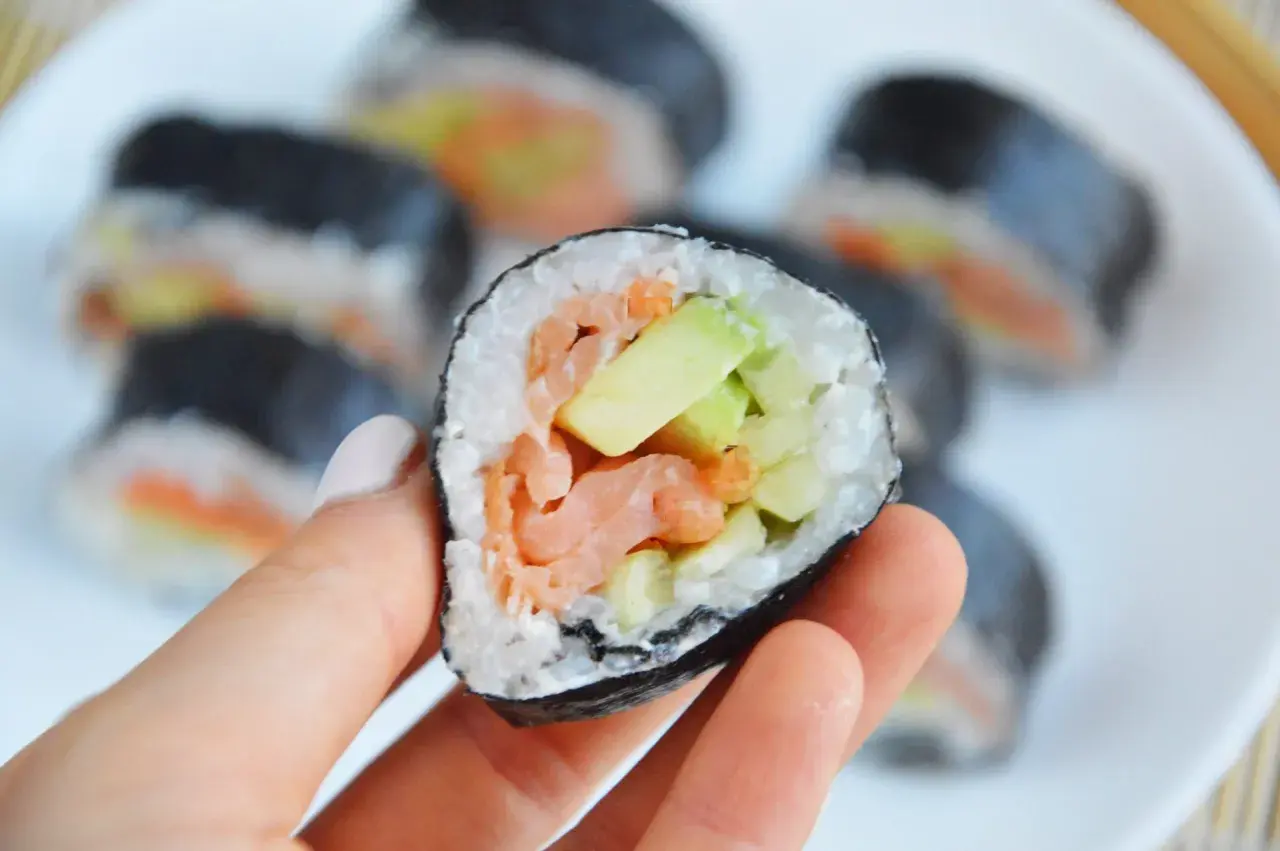 Ręka trzyma sushi z łososiem wędzonym na gorąco, awokado i ogórkiem.