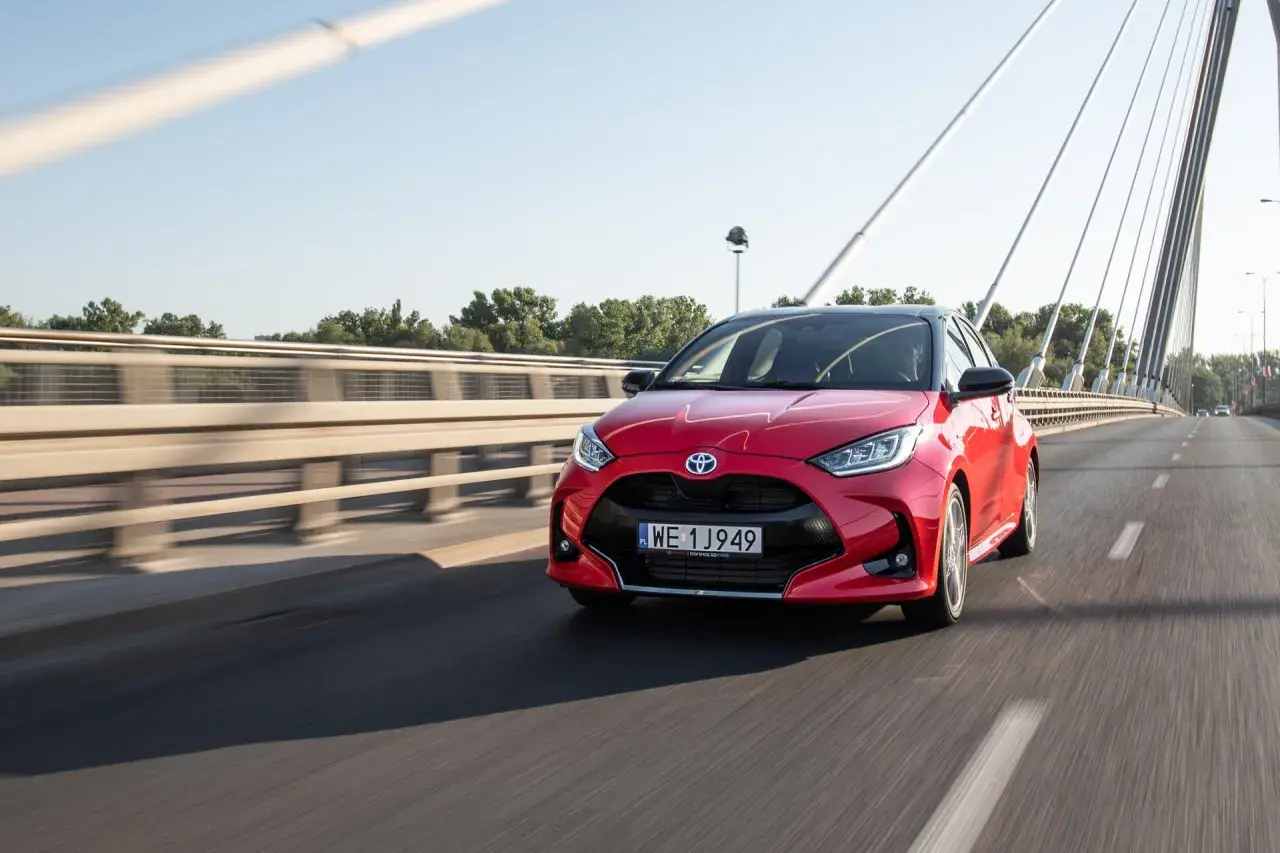 Czerwony Toyota Yaris Hybrid pędzi po moście w słoneczny dzień.