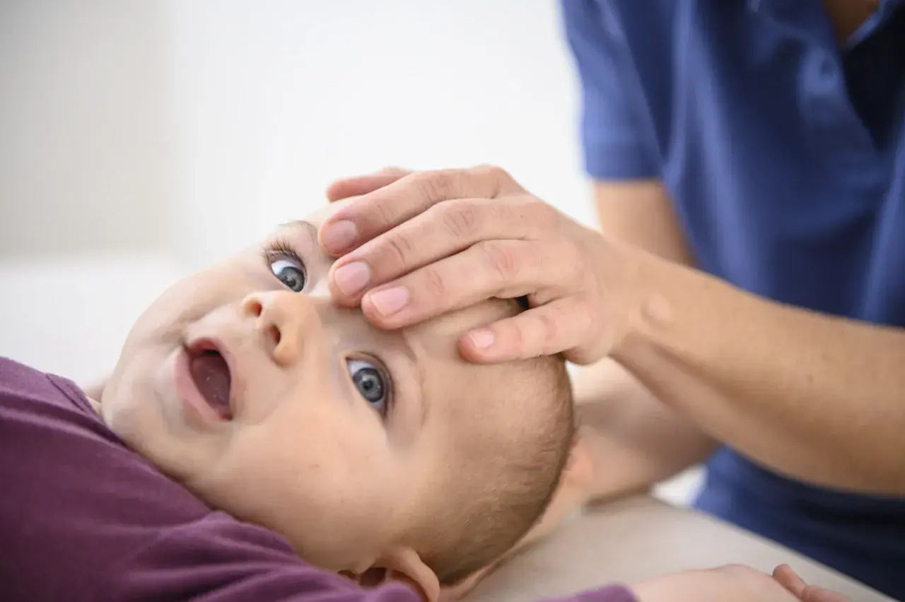 Ein Baby nach Osteopathie, wie ausgewechselt, mit weit geöffnetem Mund und großen Augen, während eine Hand sanft seine Stirn berührt.