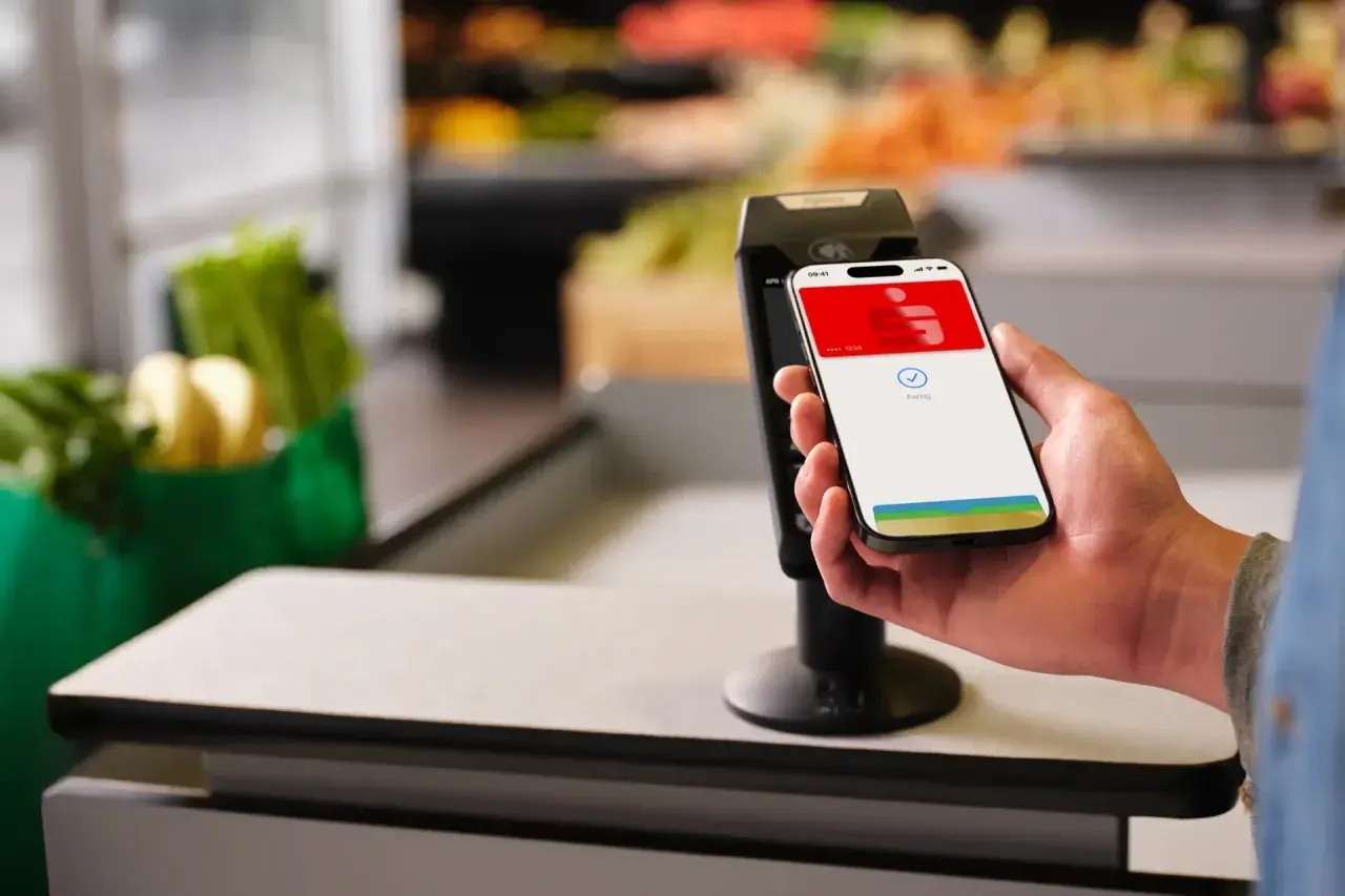 Einkaufen leicht gemacht: So funktioniert Apple Pay. Ein iPhone wird zum Bezahlen an ein Terminal gehalten, im Hintergrund sind Lebensmittel zu sehen.