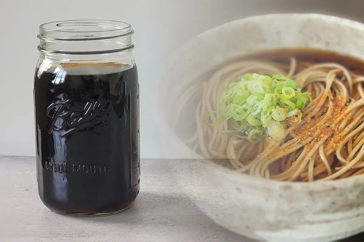 A jar of dark soba dipping sauce sits next to a bowl of soba noodles with green onions. Perfect for your soba dipping sauce recipe.