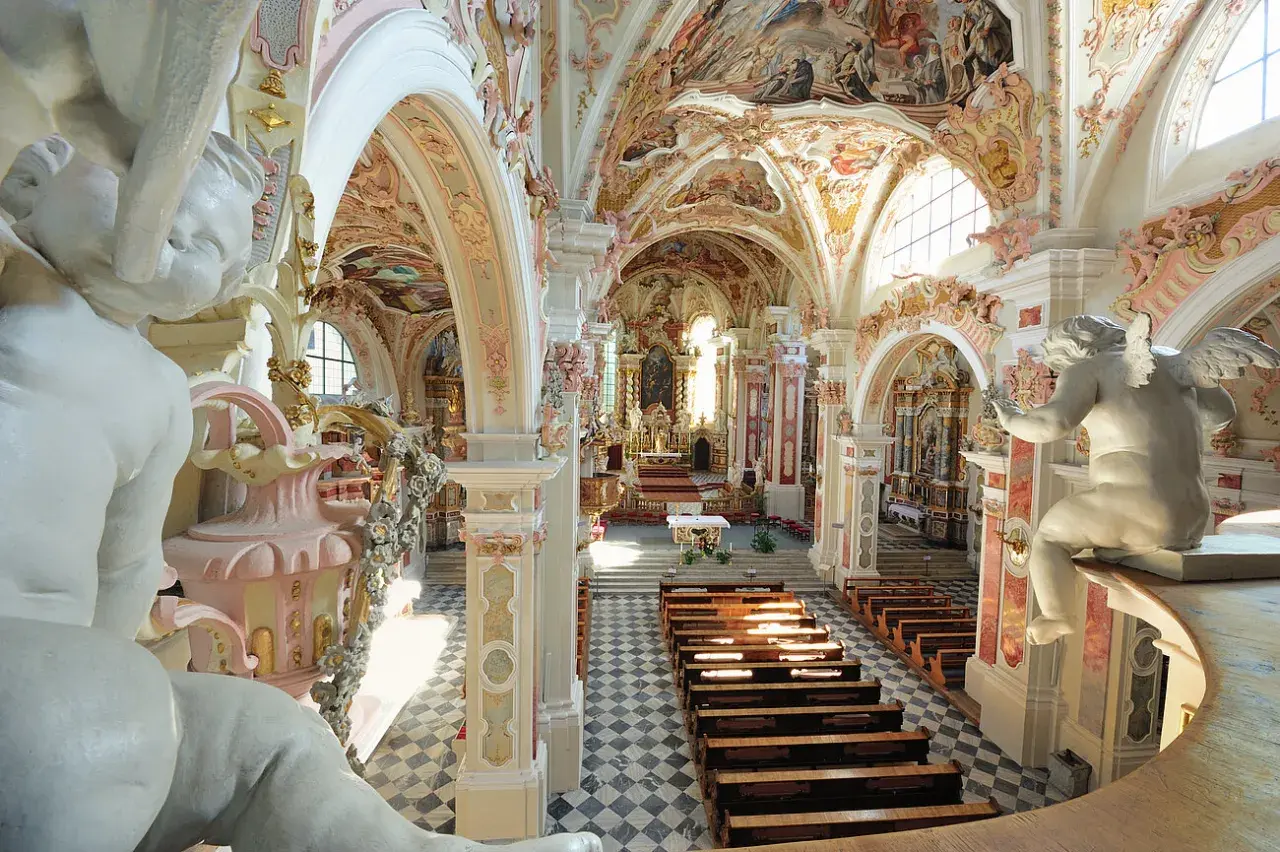 Prunkvolle Deckenmalereien und Stuckaturen im Kloster Neustift. Engelstatue blickt auf Kirchenbänke und den Altar.