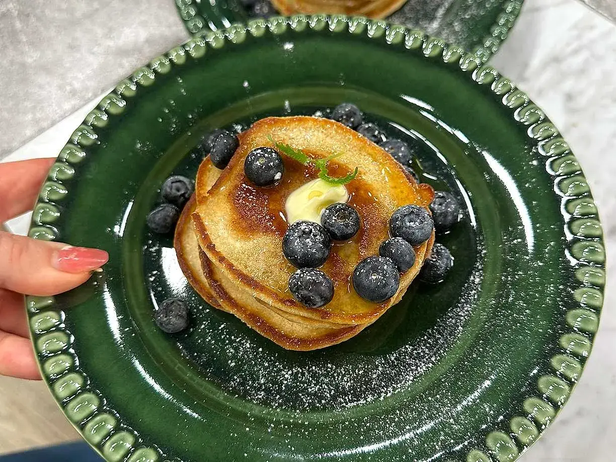 En hög med amerikanska pannkakor toppas med smör, blåbär och sirap på en grön tallrik.