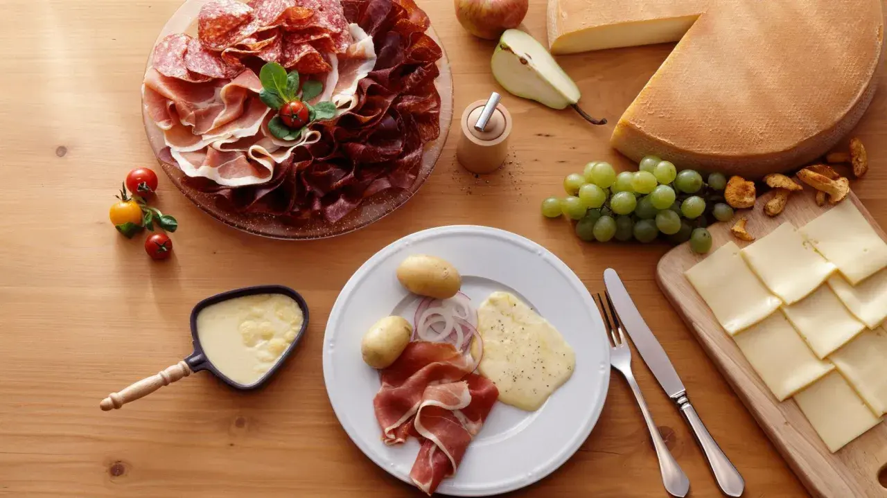 Une table dressée pour une abondance raclette : charcuteries, fromages, pommes de terre, raisins et poêlon de fromage fondu.