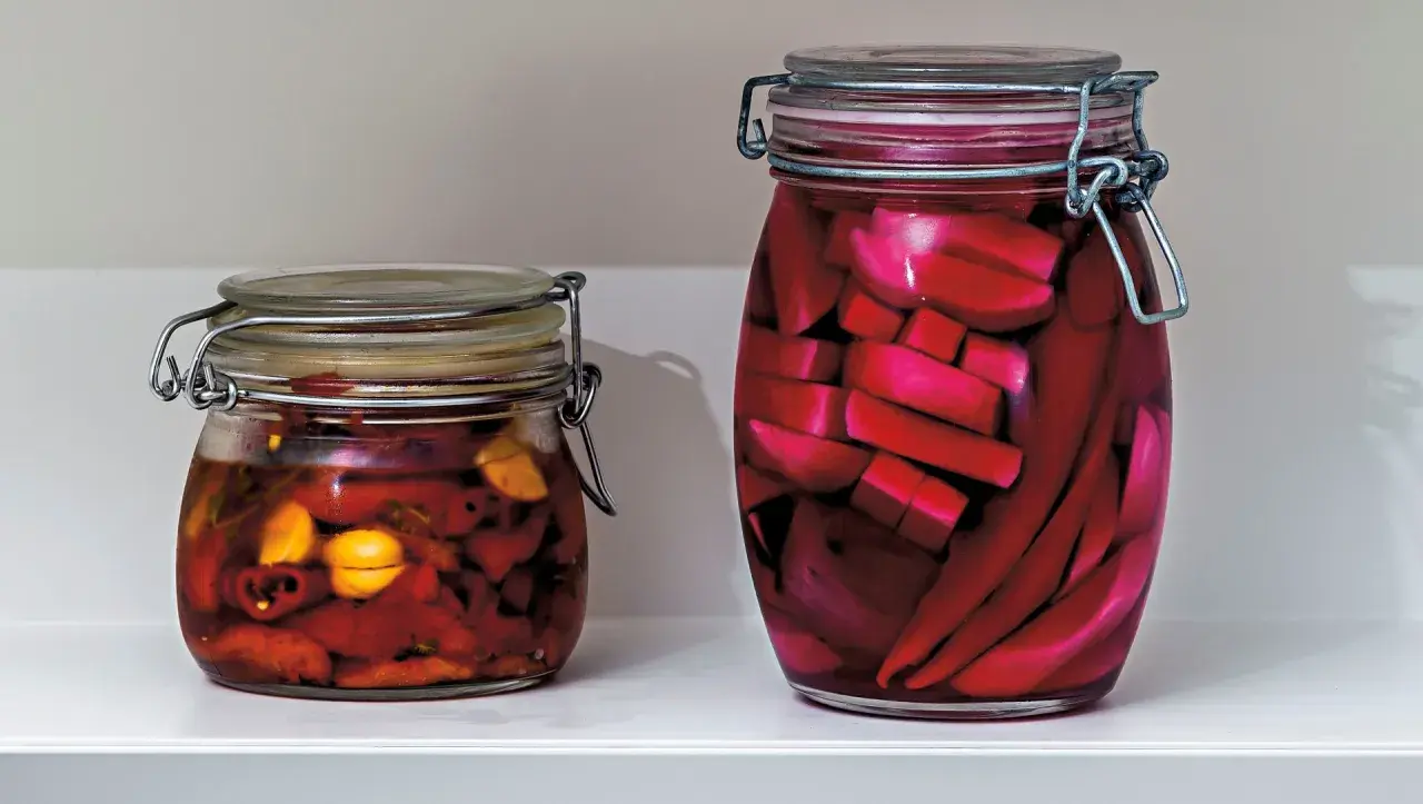 Två glasburkar med inlagd paprika och andra grönsaker. Den större burken innehåller röda, avlånga bitar, troligen inlagd paprika.