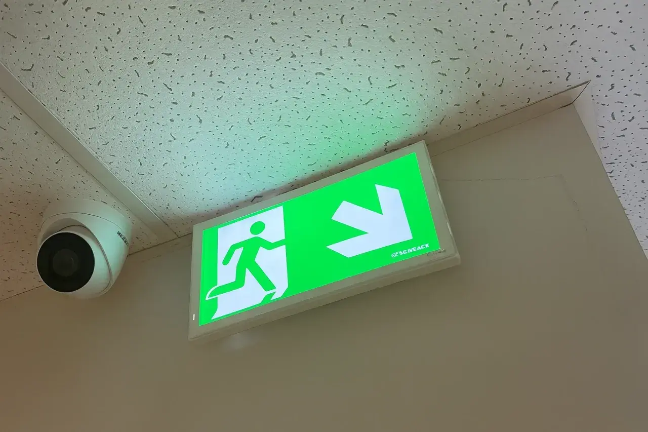 Zielone oświetlenie awaryjne z ikoną biegnącej postaci i strzałką wskazuje drogę ewakuacji. Obok widoczna kamera monitoringu.
